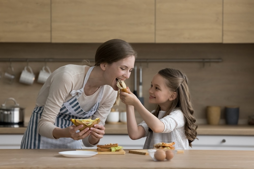 Joyful little kid girl giving mom sandwich to bite. Happy mother and daughter cooking snack for lunch together in kitchen, tasting meal, having fun, smiling, enjoying eating, homemade culinary