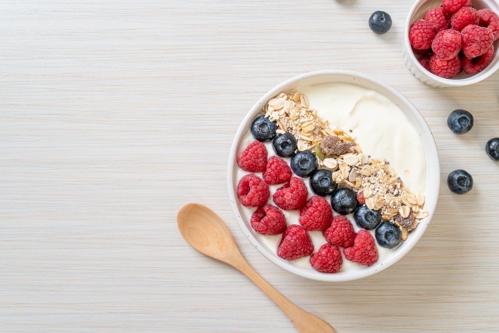 Greek yogurt, raspberries, blueberries, strawberries, granola and maple syrup photographed in ceramic bowls on a light backdrop.