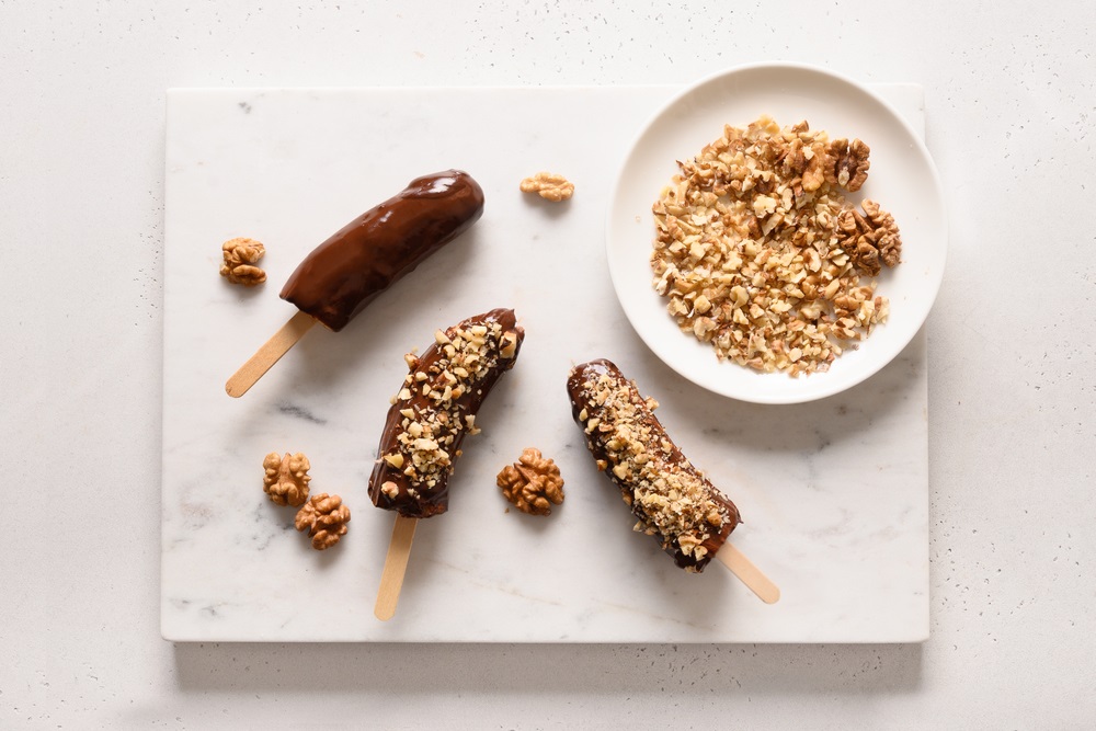 Frozen banana lollipops or lollies with walnuts sprinkles for kids on white marble table. Easy vegan sweets. View from above.